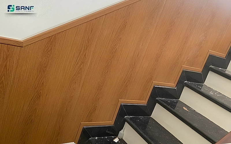 Vách tấm vân gỗ nâu cam chạy theo bậc cầu thang đá đen có nẹp trắng ở mép