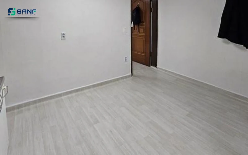 Căn phòng nhỏ lát sàn nhựa giả gỗ tông trắng xám với tường sáng và cửa gỗ