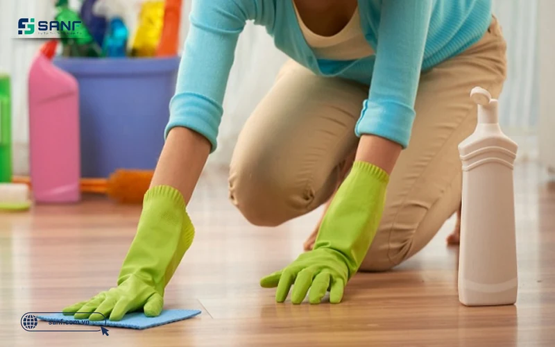 Người đeo găng tay lau sàn nhựa giả gỗ bằng khăn ẩm bên cạnh chai dung dịch vệ sinh